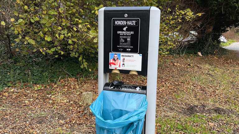 Hondenafvalstation met blauwe zak in een parkachtige omgeving.