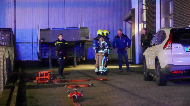 Brandweerlieden en politieagenten in uniform staan op een industrieterrein naast een geparkeerde auto en een muur.
