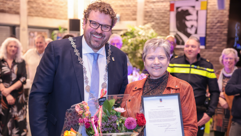 Een man en een lachende vrouw poseren met een certificaat en bloemen in een drukke zaal.