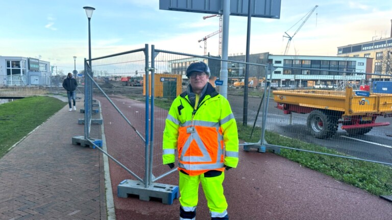 Persoon in felgele werkkleding staat op een pad naast een bouwplaats met hekwerk en machines op de achtergrond.