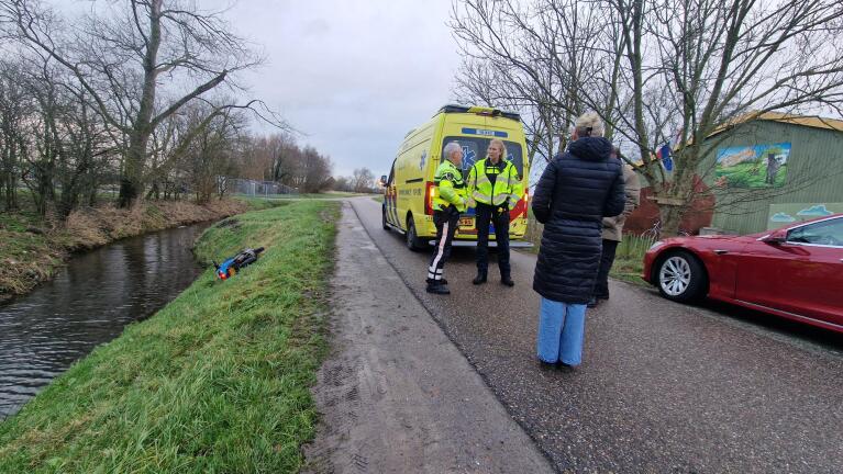 Een scooter ligt in de berm bij een sloot. Twee hulpverleners staan voor een ambulance onderweg met een vrouw in gesprek. Links zijn bomen en rechts een rode auto en een gebouw.