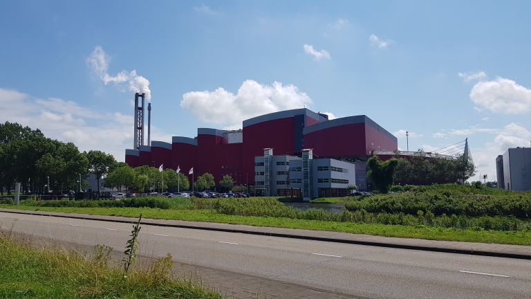 Rode industriële gebouwen met een schoorsteen, groene bomen en struiken op de voorgrond en een blauwe lucht met enkele wolken.