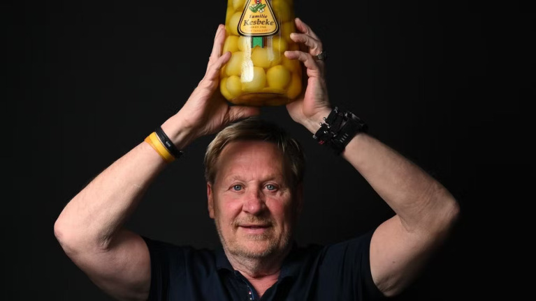 Man houdt een grote pot augurken boven zijn hoofd tegen een zwarte achtergrond.