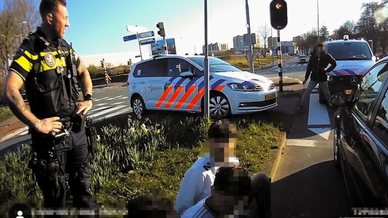 Een politieagent staat naast een politieauto bij een kruispunt met twee personen die op de grond zitten, waarvan de gezichten gepixeld zijn. Op de achtergrond is een andere politieauto zichtbaar.