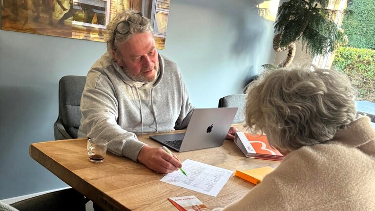 Een man zit samen met een oudere vrouw aan tafel en bespreekt documenten, met een open laptop voor zich en een glas op tafel.