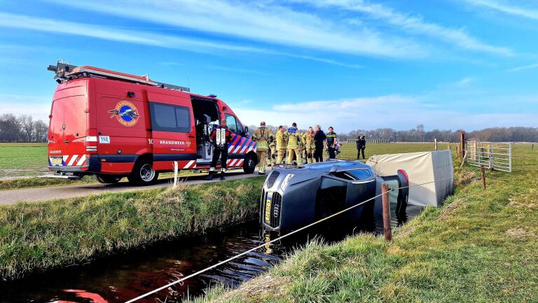 Een rode brandweerbus staat naast een greppel waar een auto op zijn kant ligt. Brandweerlieden en andere hulpverleners bekijken de situatie in een groene, landelijke omgeving.