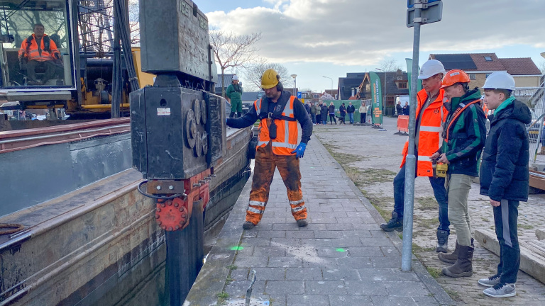 Werknemers in bouwhelmen en veiligheidshesjes werken bij een bouwplaats langs een kanaal, met een machine op de achtergrond en een paar toekijkende voorbijgangers.