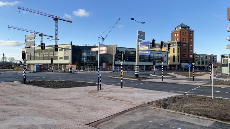 Straatbeeld van een kruispunt met verkeerslichten en wegwijzers voor een modern gebouw met de naam 'Forum'. In de achtergrond zijn bouwkranen zichtbaar en er is een rood noodtoilet geplaatst. Het weer is helder met blauwe lucht en enkele witte wolken.