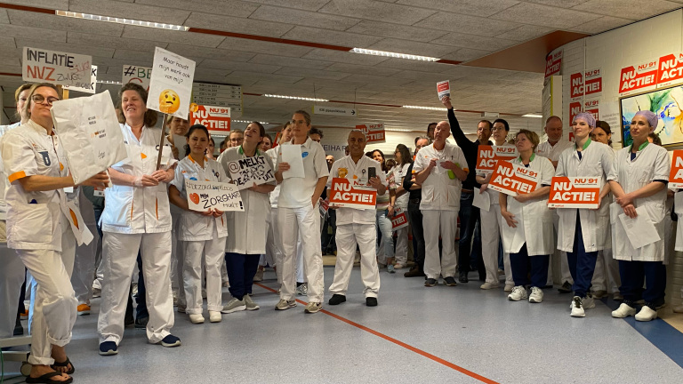 Een groep medisch personeel in witte uniformen houdt borden vast met teksten zoals 'NU ACTIE!', terwijl ze in een ziekenhuisgang staan. Ze lijken te protesteren of actie te voeren tegen inflatie en voor betere arbeidsomstandigheden. De omgeving is een ziekenhuisgang met posters en instructieborden op de muren.