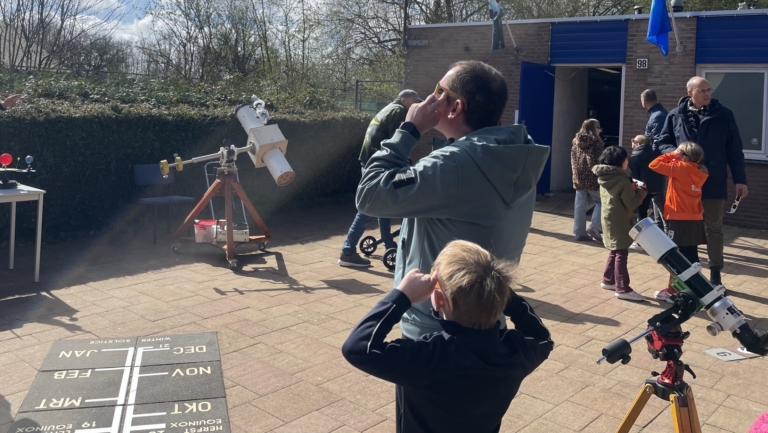 Groep mensen verzameld op een zonnige dag, sommige kijken door telescopen terwijl anderen naar de lucht staren met speciale brillen, omringd door bomen en een gebouw met blauwe deuren.