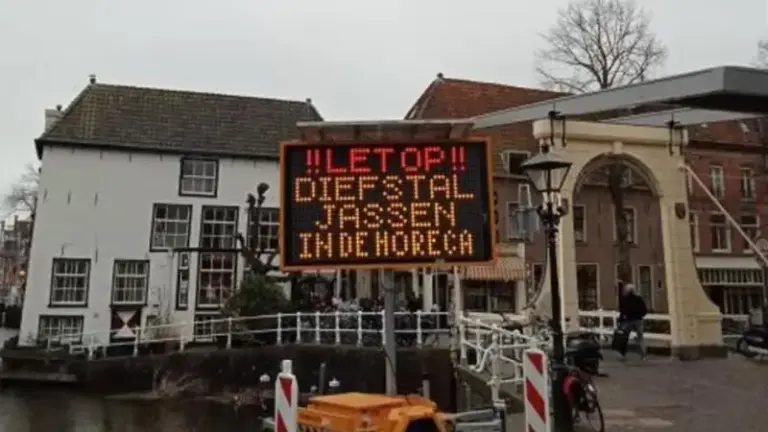 Elektronisch verkeersbord met de tekst: 'LET OP!! DIEFSTAL JASSEN IN DE HORECA', geplaatst bij een brug in een historische wijk met bakstenen gebouwen.