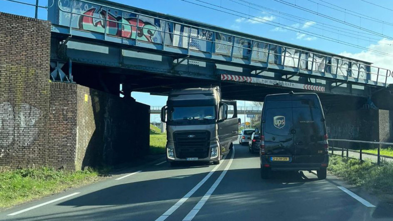 Een vrachtwagen zit vast onder een lage brug, terwijl een UPS-busje ervoor staat op de weg. Boven de brug is graffiti zichtbaar.