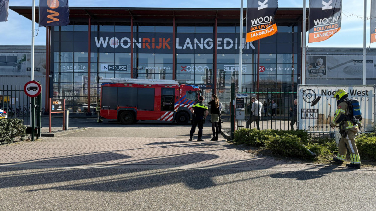 Een brandweerwagen geparkeerd voor het Woonrijk Langedijk pand, met een politieagent en een vrouw die met elkaar praten, terwijl een brandweerman in uniform op de voorgrond staat.