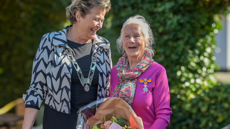 Twee vrouwen staan naast elkaar buiten, lachend. De vrouw rechts draagt een roze trui en een kleurrijke sjaal, met een medaille op haar borst en een boeket bloemen in haar handen. De vrouw links draagt een ketting met een medaille en heeft een zwart-witte zigzag blazer aan.