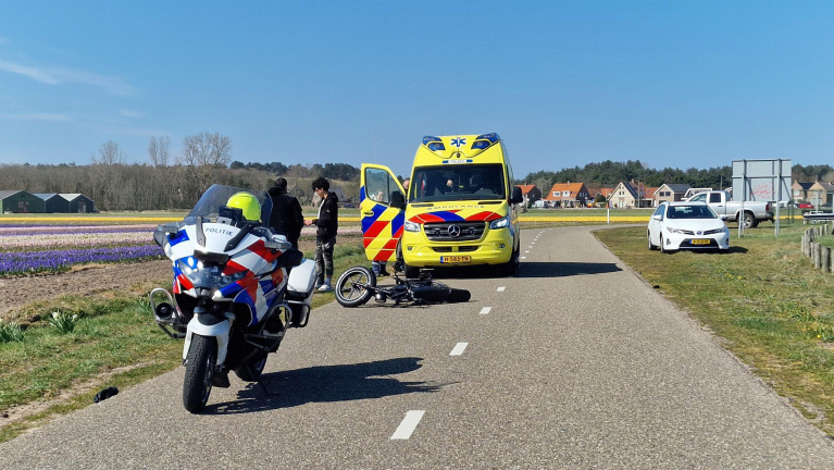 Een Nederlandse ambulance en een politiemotor staan op een landweg in de buurt van een omgevallen fiets. Er zijn mensen ter plaatse, en een dorp en bloemenveld zijn te zien op de achtergrond.
