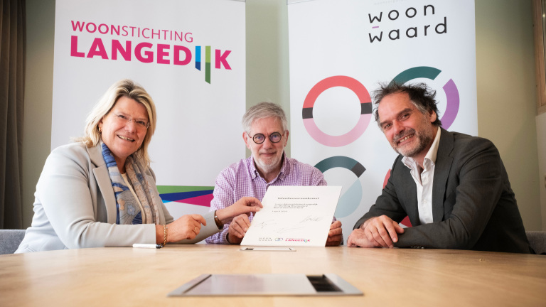 Drie mensen poseren met een ondertekend document aan een tafel, met op de achtergrond banners van "Woonstichting Langedijk" en "Woonwaard".
