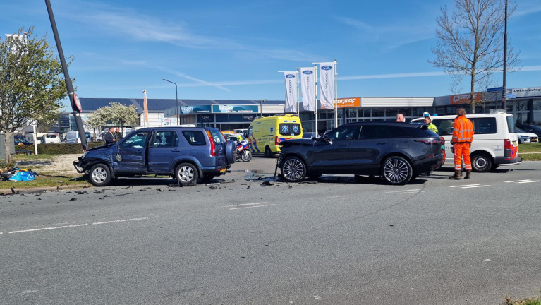 Twee auto's na een aanrijding op een wegencomplex, omringd door hulpverleners en een ambulance op de achtergrond.