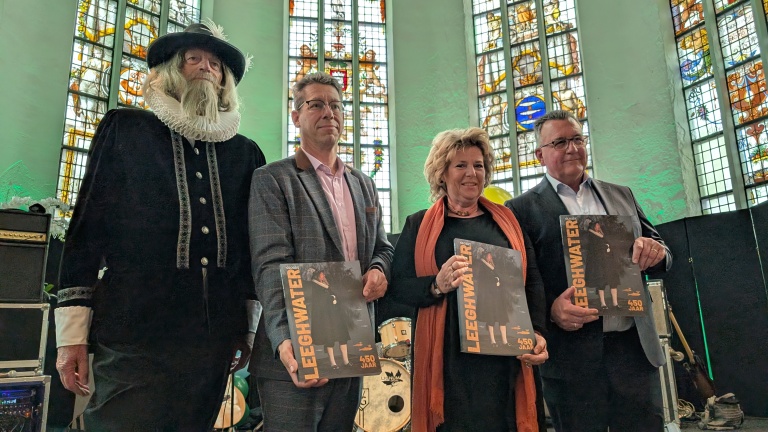 Vier mensen poseren met tijdschriften in hun handen, terwijl ze in een ruimte met grote, kleurrijke glas-in-loodramen staan.