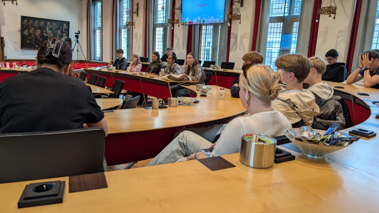Een groep mensen zit in een kring in een vergaderzaal met een rond tafelblad en kijken naar een presentatie op een scherm waarop 'Vragen?' staat. De zaal heeft grote ramen en een schilderij aan de muur, en er staan merken van drankjes en snacks op de tafel.
