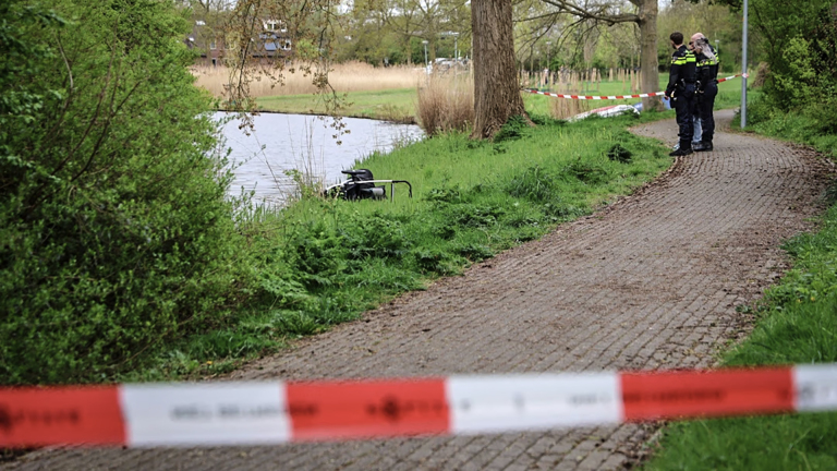 Twee politiemensen staan op een met rood-wit lint afgezet pad langs een vijver, omringd door bomen en struiken.