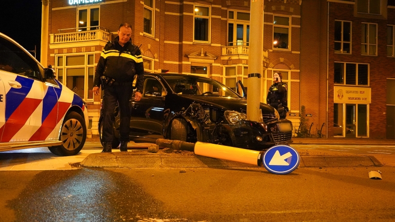 Een politieagent bij een gecrashte zwarte auto die tegen een paal botste, met een omgevallen verkeersbord op de grond naast de auto in een stedelijke omgeving 's nachts.
