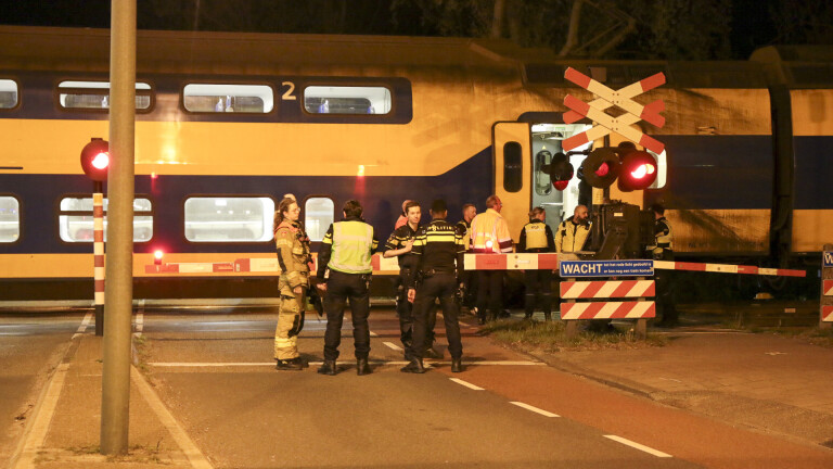 Groep politieagenten en hulpverleners staan bij een spoorwegkruising naast een stilstaande trein met rode waarschuwingslichten, tijdens de nacht.