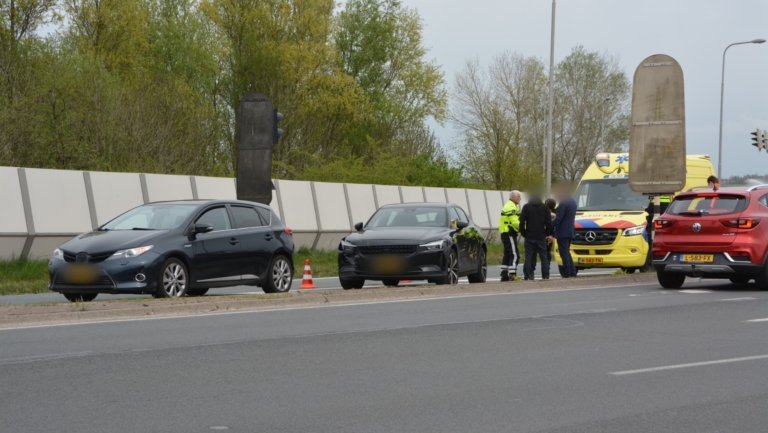 Twee auto's en een ambulance staan stil langs een weg met kegels, terwijl mensen in gesprek zijn. Op de achtergrond zijn bomen en een geluidsscherm zichtbaar.