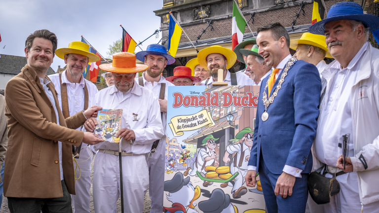 Een groep mannen, waaronder enkele in traditionele kaasdragerskleding met kleurrijke hoeden, poseert buiten met een groot Donald Duck tijdschrift. Een man in een blauw pak en een ketting lacht. Op de achtergrond zijn vlaggen te zien.