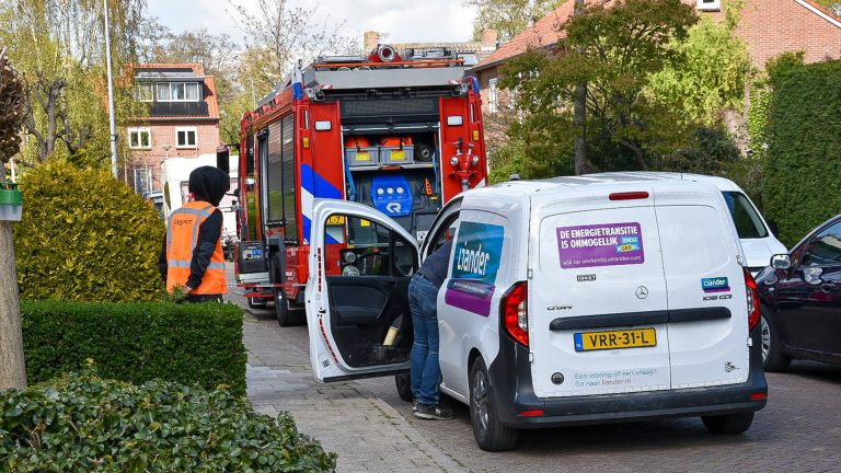 Een witte bestelwagen met het logo van Liander en een sticker over energietransitie staat geparkeerd naast een rode brandweerwagen op een straat. Een persoon in een oranje veiligheidsvest en donkere kleding loopt langs de auto. Er zijn bomen en een huis op de achtergrond.