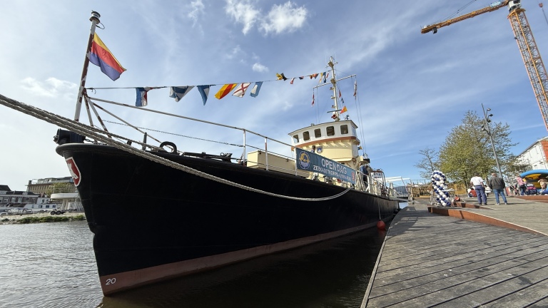 Een zwart schip met kleurrijke vlaggen, een bord met 'Open Dag' en enkele mensen op de kade bij de haven, omgeven door een kraan en gebouwen.