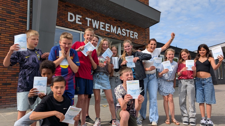 Een groep lachende kinderen en een volwassene staan buiten voor een gebouw genaamd 'De Tweemaster', terwijl ze papieren vasthouden.