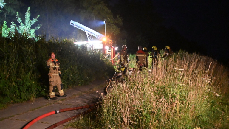 Brandweerlieden in uniform staan 's nachts op een pad omgeven door hoge grasvelden, met een brandweerwagen op de achtergrond.