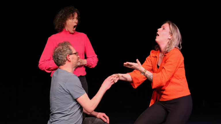 Drie mensen op een podium; een zittende man met een bril in een grijs T-shirt reikt naar een vrouw in een oranje jasje die een verbaasde uitdrukking heeft, terwijl een vrouw in een roze trui achter hen met een geschokte blik toekijkt.