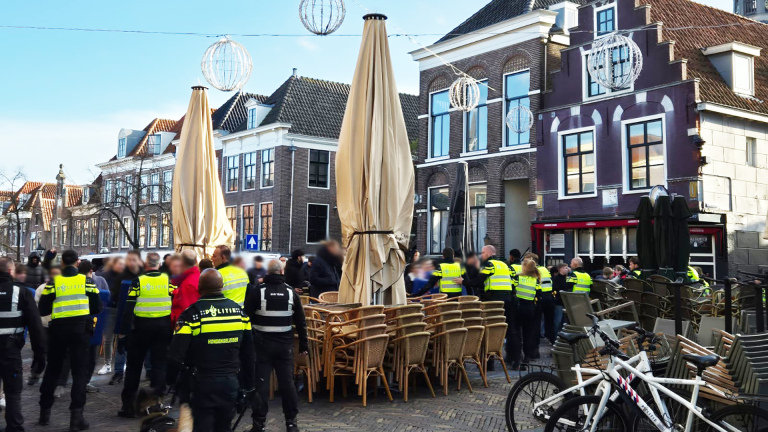 Groep politieagenten in gele hesjes staat op een plein met opgestapelde terrasstoelen voor oude gebouwen in Nederland.
