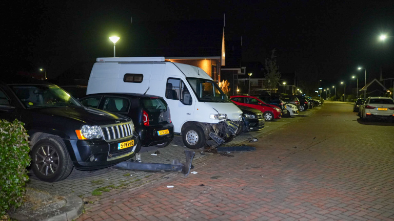 Een nachtelijke scène van een botsing tussen een witte bestelwagen en een zwarte auto op een parkeerplaats. De voorkant van de bestelwagen is beschadigd en er ligt wrak op de grond rondom de voertuigen.