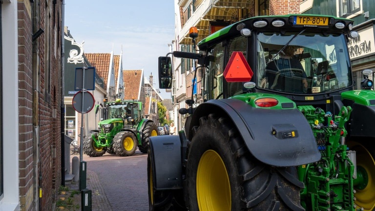 Twee groene tractoren rijden door een smalle stadsstraat met bakstenen gebouwen aan weerszijden.