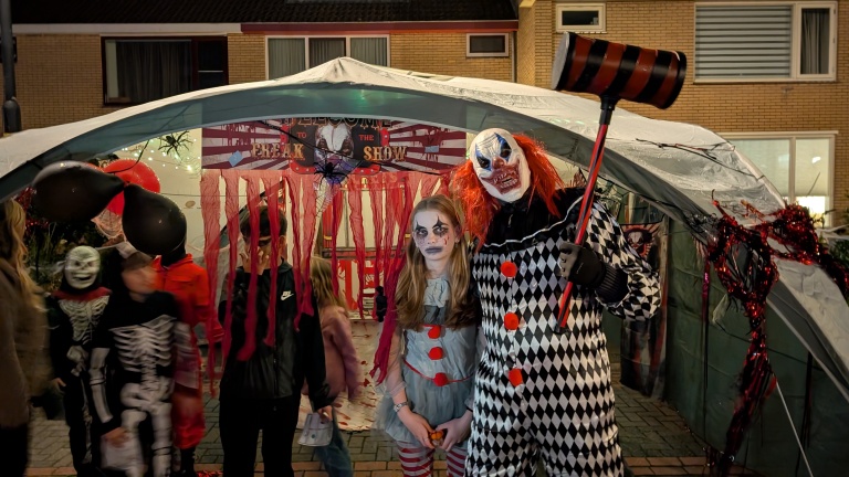 Mensen verkleed als enge clowns en skeletten tijdens een Halloween-evenement, poseren voor een tent met een spandoek waarop 'Welkom bij de Freak Show' staat.