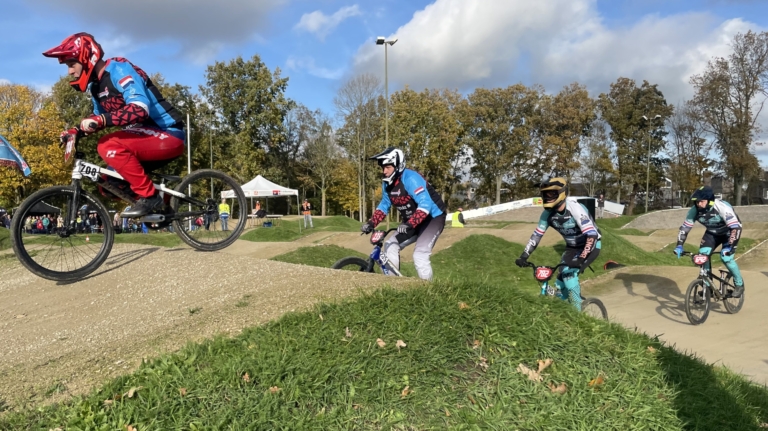 BMX-fietsers in felgekleurde pakken racen over een heuvelachtig parcours met grasrijke delen en een heldere blauwe hemel op de achtergrond.