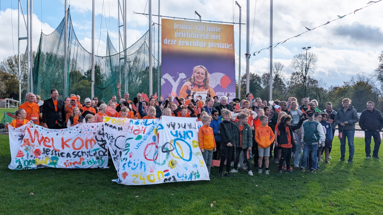 Een groep mensen, waaronder veel kinderen in oranje kleding, poseert met spandoeken op een grasveld. Op de achtergrond is een groot billboard te zien met een felicitatietekst voor Jessica en een afbeelding van een vrouw met een medaille.
