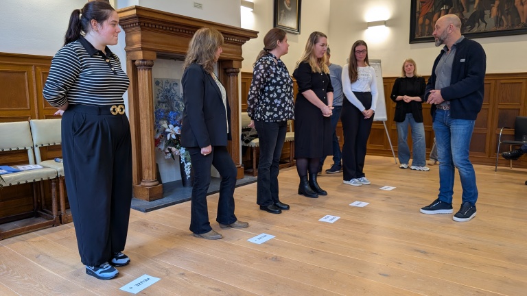 Groep mensen staat in een kamer met houten vloer en een haard, kijkend naar een man die voor een instructeur lijkt te staan. Op de vloer liggen papieren met woorden erop.