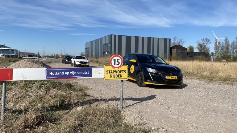 Twee auto's rijden op een onverharde weg langs een bord met de tekst "Holland op zijn smalst" en een ander bord dat een snelheidslimiet van 15 aangeeft met "stapvoets rijden." Op de achtergrond staan gebouwen en een windmolen.