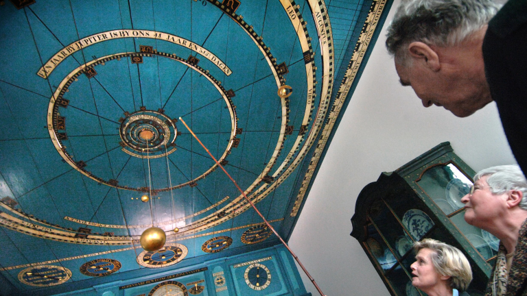 Mensen kijken omhoog naar een model van een planetarium op een blauwe plafond met cirkels en sterrenbeelden, met een kast met borden op de achtergrond.