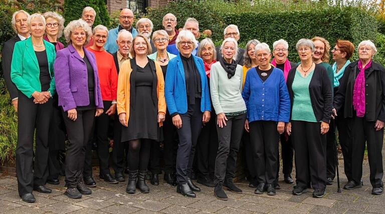 Groepsfoto van een koor bestaande uit mannen en vrouwen van middelbare tot oudere leeftijd, staande buiten voor groene struiken. Ze dragen kleurrijke kleding, waaronder jasjes in blauw, groen, oranje, en paars.