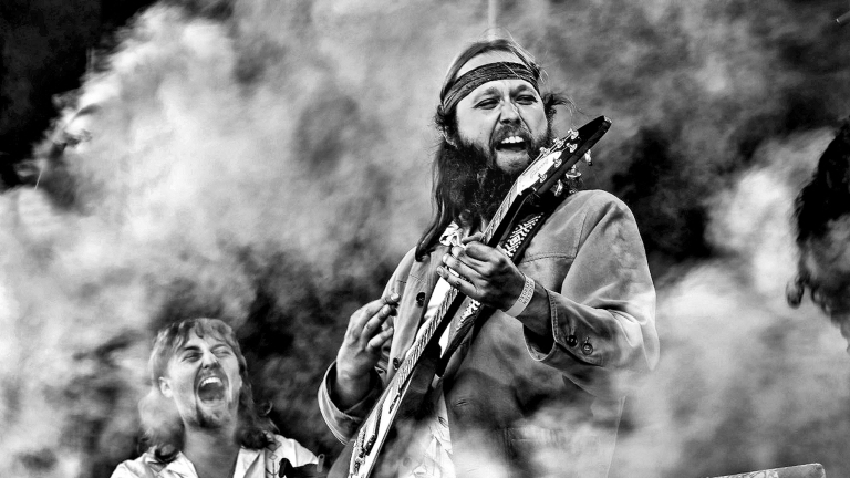 Twee muzikanten in een zwart-witfoto; de man op de voorgrond speelt gitaar en draagt een hoofdband, terwijl de andere man lacht, omringd door rook of mist.