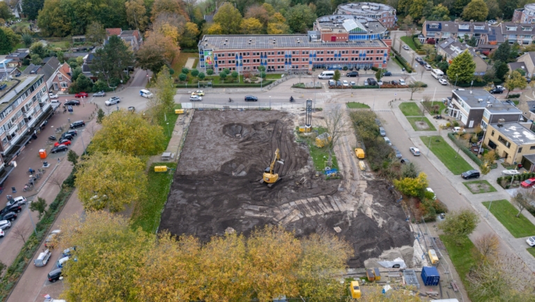 Luchtfoto van een bouwplaats omgeven door bomen en gebouwen, met een graafmachine aan het werk op een braakliggend terrein en verschillende geparkeerde voertuigen eromheen.