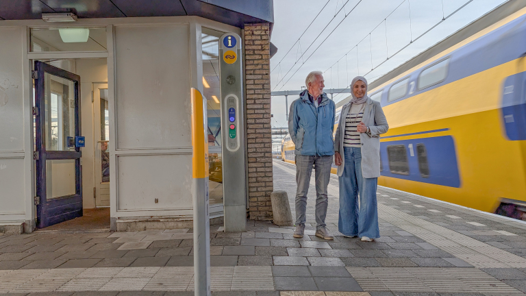 Twee mensen staan op een treinstation met een gele en blauwe trein die op de achtergrond voorbijrijdt.