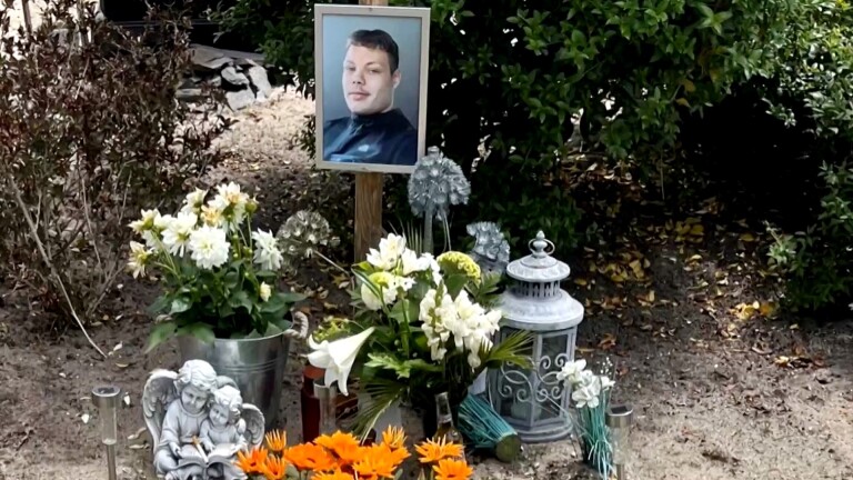 Een herdenkingsplek met een portretfoto van een man, omringd door verschillende bloemen in potten, een decoratief lantaarn, en een beeldje van een engel op een boek lezend kind. De bloemen zijn voornamelijk wit en oranje, en de omgeving is groen met struiken.