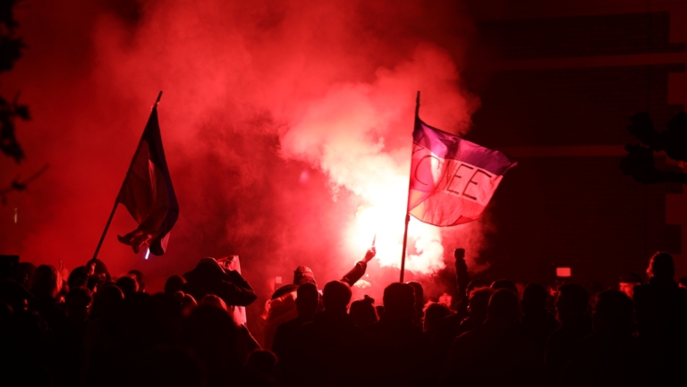 Een menigte mensen met vlaggen en fakkels staat in het donker, verlicht door een felrode gloed van vuurwerk.