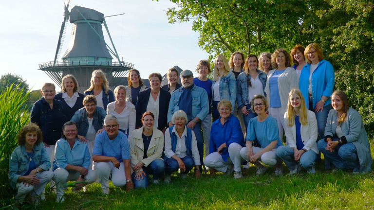 Een groep mensen poseert in het gras voor een traditionele Nederlandse windmolen, omringd door groen en een heldere lucht.