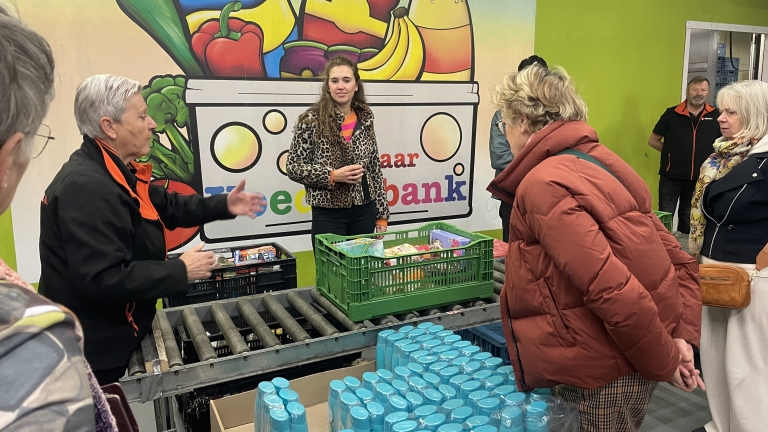 Een groep mensen staat rond een tafel in een voedselbank met kratten gevuld met voedsel en een muur met een kleurrijk schilderij van groenten en fruit op de achtergrond.